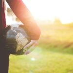 person holding black and brown globe ball while standing on grass land golden hour photography